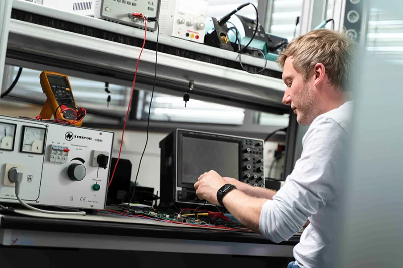 Person mit weißem Langarmshirt bedient ein Messgerät an einem Labortisch mit mehreren elektronischen Geräten und Kabeln.